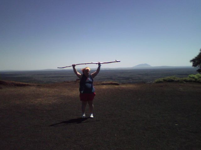 Person holding a long stick overhead on a dark, barren landscape with distant hills under a clear blue sky