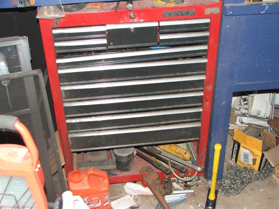 Red Craftsman tool chest with black drawers, next to a blue workbench, with tools and supplies scattered around