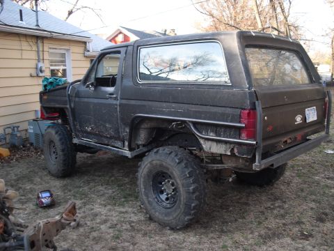 1986 Ford Bronco - This modified black SUV features large off-road tires and a custom roll cage, seen from the rear three-quarter angle.