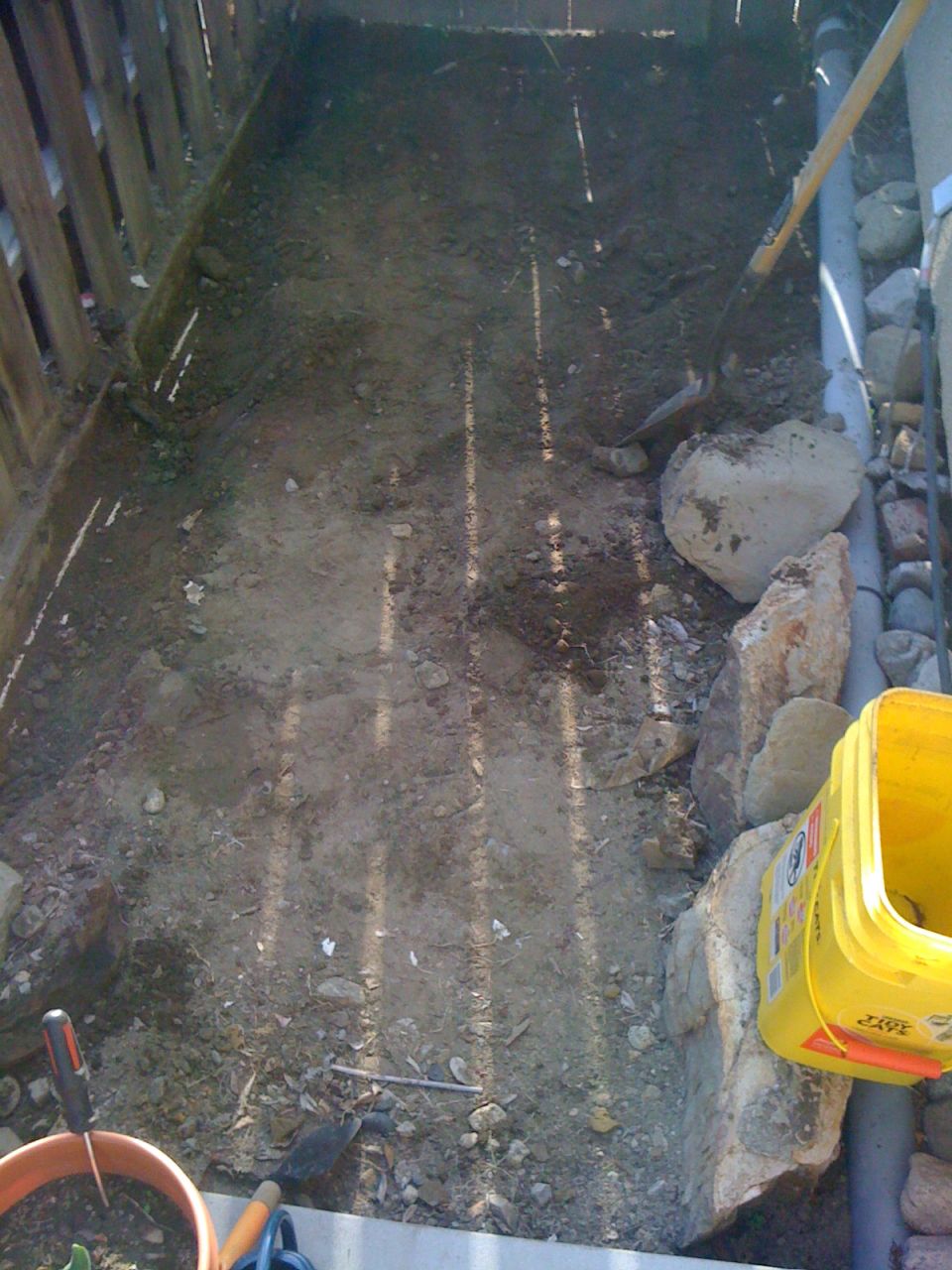 dirt area next to a wooden fence, with a shovel, rake, and a yellow bucket
