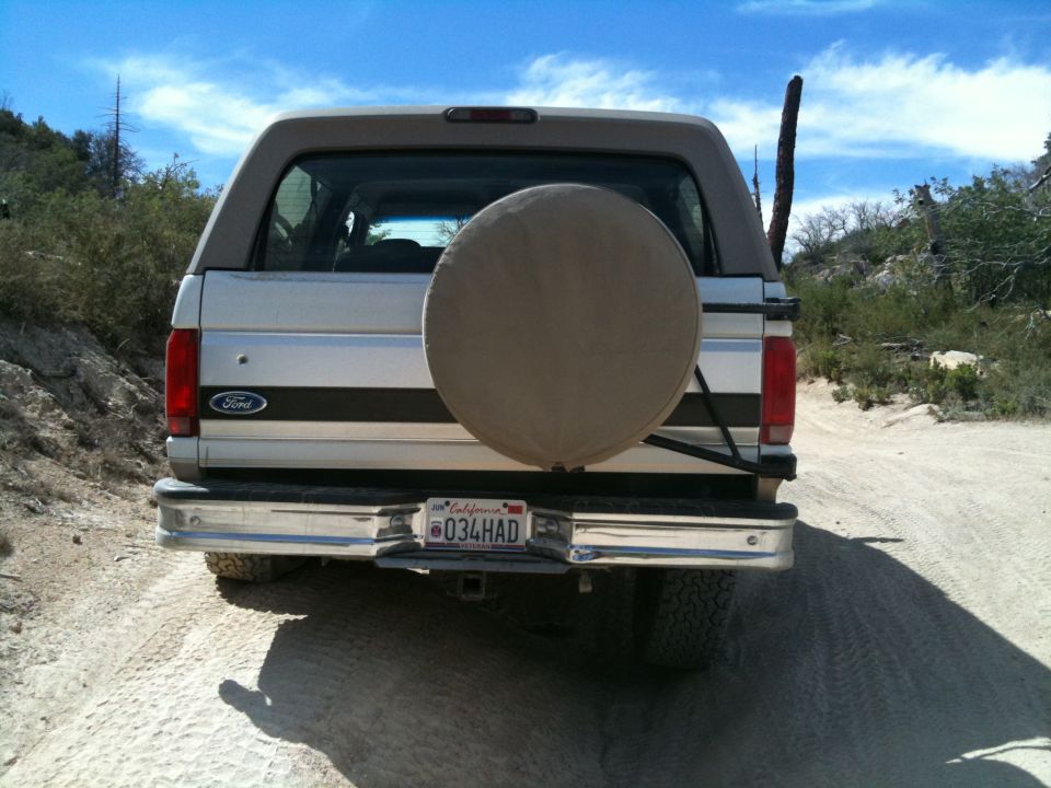 Rear of tan Ford Bronco SUV with spare tire cover and California plate on a dirt trail.