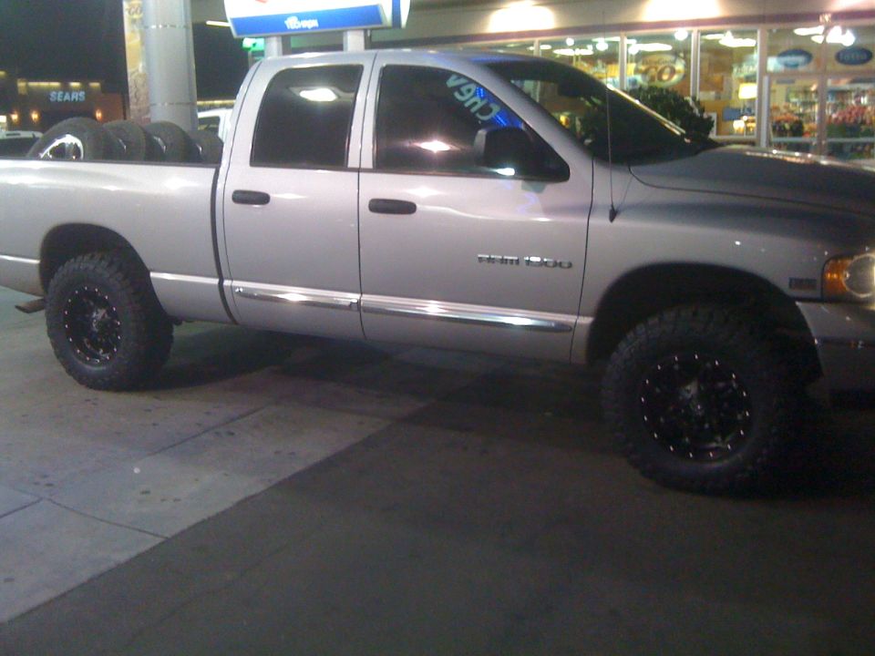 Silver Dodge Ram 1500 crew cab pickup, side view, black wheels, parked at a gas station at night.