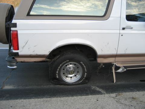 1996 Ford Bronco - Flat tire on a 1996 Ford Bronco, showing the rear driver's side wheel and body damage.