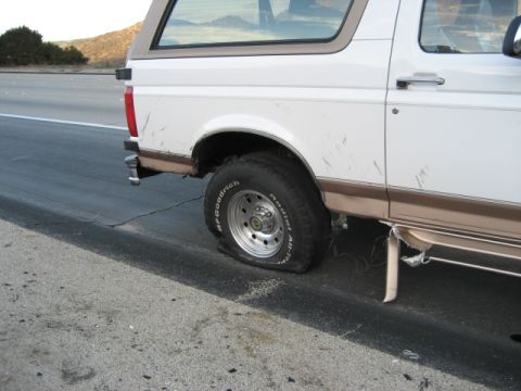 1996 Ford Bronco - The rear tire of a white Ford Bronco is completely flat while parked on the side of a highway.