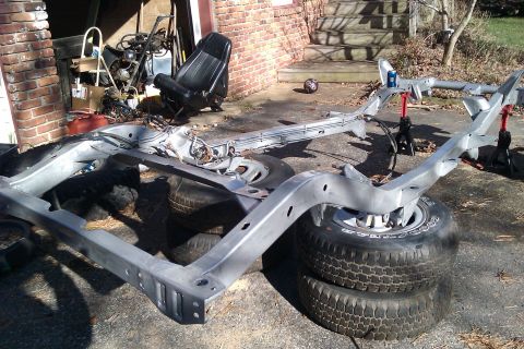 1989 Jeep Wrangler - The bare frame of a vehicle is supported by jack stands, with spare tires and a car seat visible.