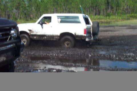 1995 Ford Bronco - Side view of a mud-splattered white full-size Ford Bronco churning through a muddy bog pit, a spare tire mounted at the rear and a tall ant…