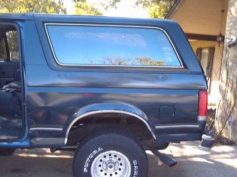 1990 Ford Bronco - Passenger-side rear view of a dark teal/blue Ford Bronco with the removable hardtop in place, showing the large fixed side glass, fuel door…