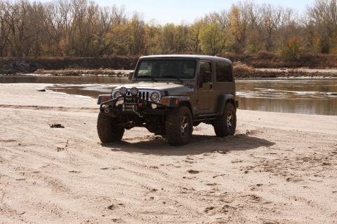 2005 Jeep Wrangler - A modified Jeep Wrangler sits on a sandy shore next to a calm river under a clear sky.