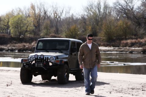 2005 Jeep Wrangler - A man walks away from a gray Jeep Wrangler parked on a sandy riverbank with trees in the background.