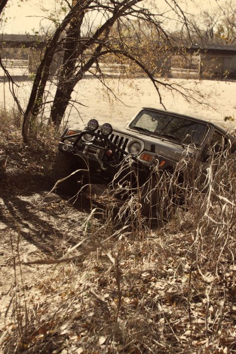 2005 Jeep Wrangler - A 2005 Jeep Wrangler is partially submerged in mud and vegetation, with trees and a bridge in the background.