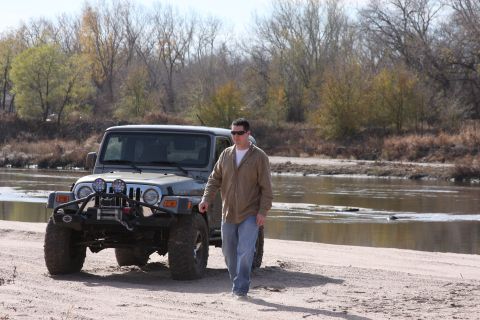 2005 Jeep Wrangler - A man walks beside a gray Jeep Wrangler parked on a sandy riverbank with trees and water in the background.
