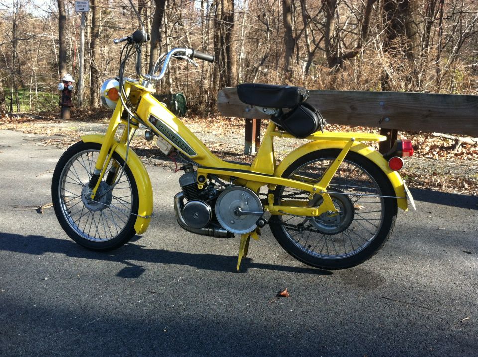 Yellow moped with black seat parked on asphalt, trees in background