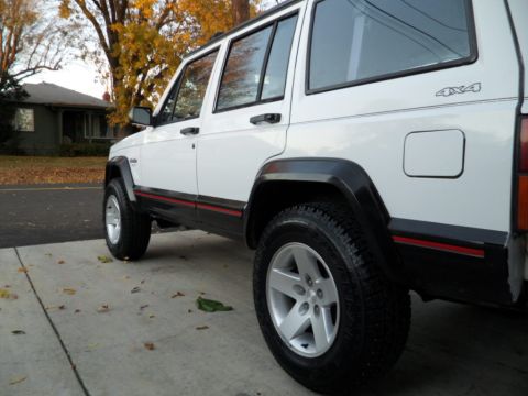 1994 Jeep Cherokee - A white Jeep Cherokee is seen from the rear three-quarter view, parked on a concrete driveway with autumn leaves scattered around.