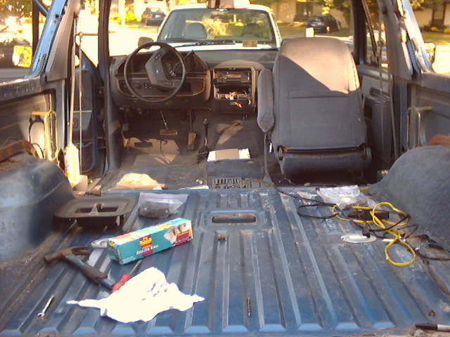 1990 Ford Bronco interior, stripped down with seats removed and dashboard exposed. Another vehicle is visible through the windshield