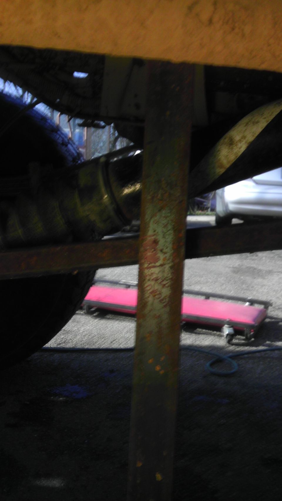 Underside view of a vehicle's suspension components and exhaust pipe. A pink object on wheels is visible in the background
