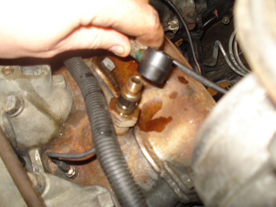 Hand holding a tool on a brass fitting screwed into a rusty engine component, with black wiring loom nearby