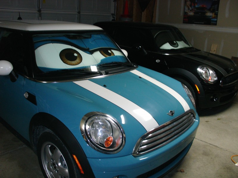 Blue MINI Cooper with white bonnet stripes and cartoon-eye sunshade beside a black MINI in a garage