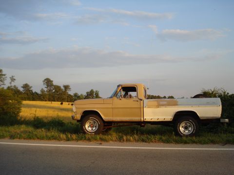 1967 Ford F-250 - A two-tone Ford F-Series truck sits parked on the shoulder of a road, with a grassy field and trees in the background.
