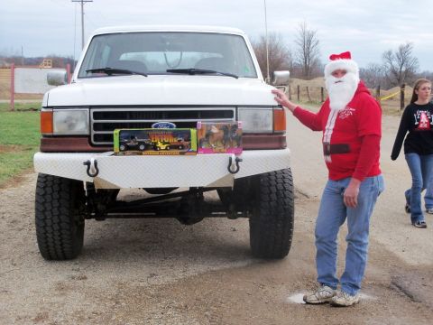 1989 Ford Bronco - A white Ford truck is parked with toys on its bumper, while a man dressed as Santa Claus stands beside it.