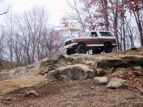 1989 Ford Bronco - A two-tone Ford Bronco navigates a rocky off-road course in a wooded area.