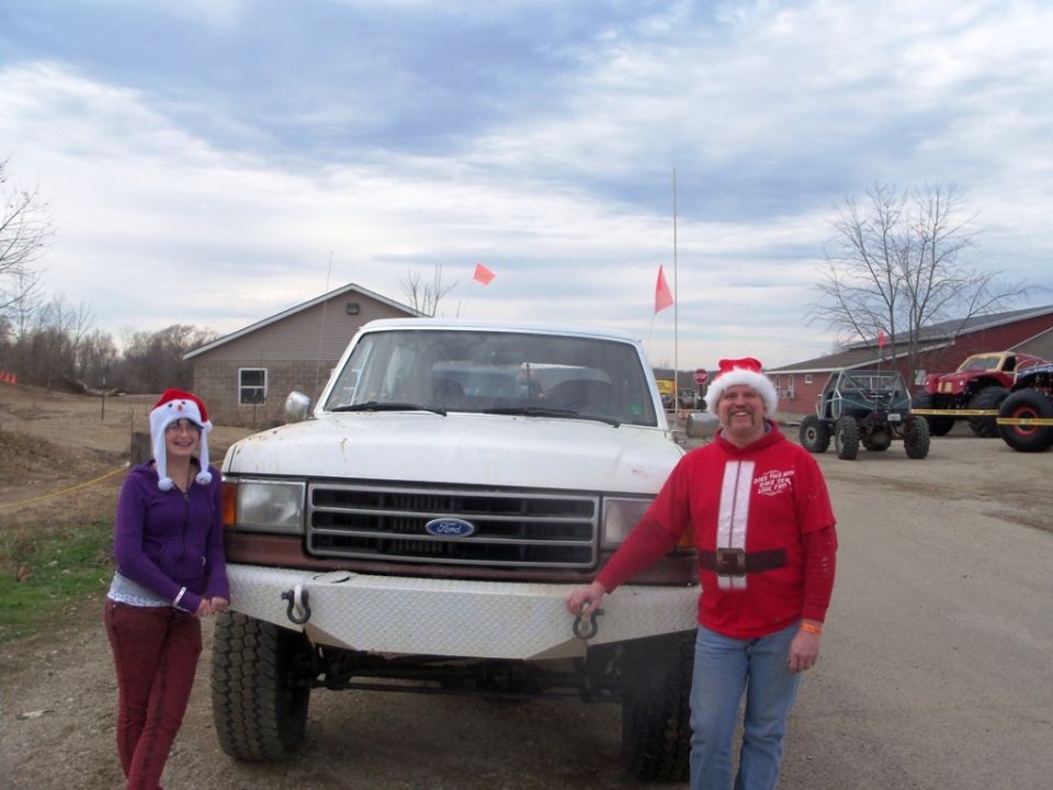 1989 Ford Bronco with a custom bumper, two people in Santa hats standing nearby. Other off-road vehicles are visible in the background