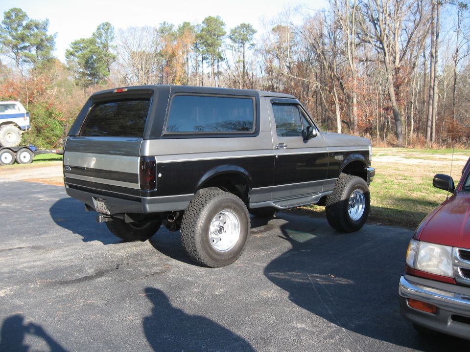 Lifted black and gray Ford Bronco parked on asphalt, red truck to the right, another vehicle on a trailer in the background