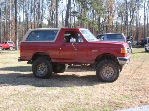 1994 Ford Bronco - Lifted red Ford Bronco with a tan removable top is parked outdoors with other vehicles nearby.