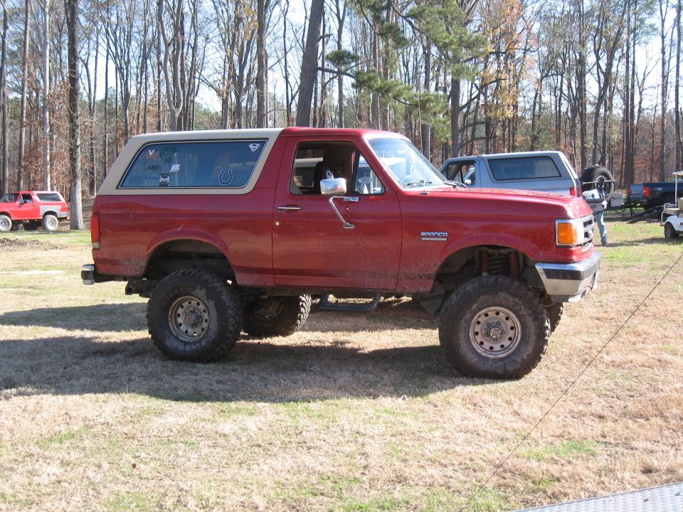 Red lifted Ford Bronco parked on grass, tan top. Another SUV visible behind it