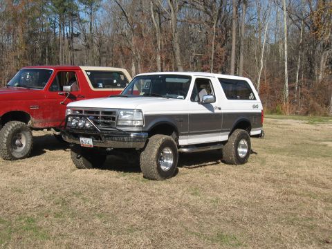 1994 Ford Bronco - Two lifted SUVs, a red one and a white and grey one, are parked on a grassy field with trees in the background.