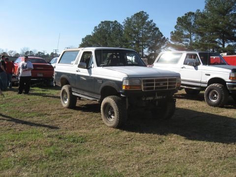 1994 Ford Bronco - Two lifted trucks, a Ford Bronco and a Ford F-Series, sit parked outdoors on a sunny day.