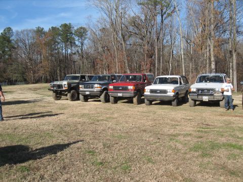 1994 Ford Bronco - Multiple Ford Bronco SUVs are lined up in a field on a clear day, with bare trees in the background.