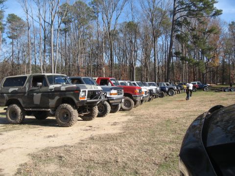 1994 Ford Bronco - A row of off-road vehicles, including a camouflaged Ford Bronco, are parked on a grassy field near a wooded area.