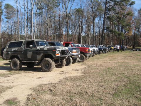 1994 Ford Bronco - A line of off-road vehicles, including a camouflaged SUV, are parked outdoors on a dirt and grass lot.