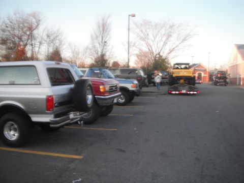 1989 Ford Bronco - Several vehicles are parked in a lot, including a silver Bronco and a red pickup truck, with a tow truck and trailer visible further back.