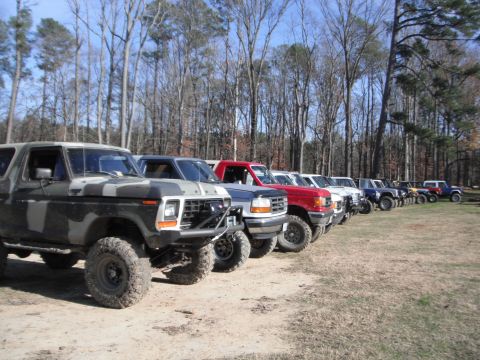 1989 Ford Bronco - Multiple off-road vehicles are lined up in a dirt lot with trees in the background.