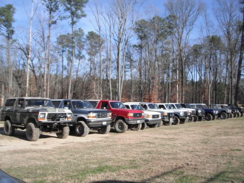 1989 Ford Bronco - A row of Ford Broncos and F-Series trucks are parked in a grassy field with a line of trees in the background.