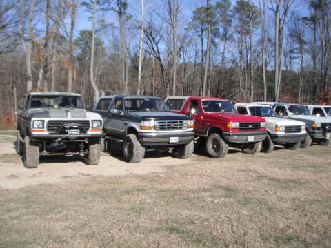 1989 Ford Bronco - A lineup of modified Ford trucks and SUVs, including an early model Bronco, are parked outdoors on a dry, grassy field.