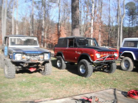 1989 Ford Bronco - Three Ford Broncos are parked outdoors on a grassy area with trees in the background. The middle vehicle is red with a black hood and soft…
