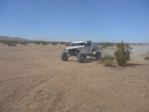 1988 Ford Bronco - A custom off-road buggy navigates a dusty desert terrain under a clear blue sky, with another vehicle visible in the distance.