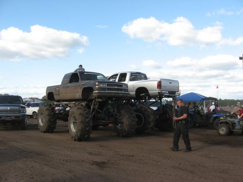 1994 Ford Bronco - Monster trucks with pickup truck bodies are shown at an outdoor event with other vehicles and people.