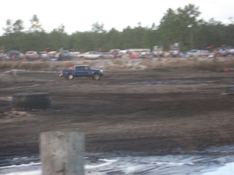 1994 Ford Bronco - A blue pickup truck navigates a muddy course with spectators and parked vehicles visible in the distance.