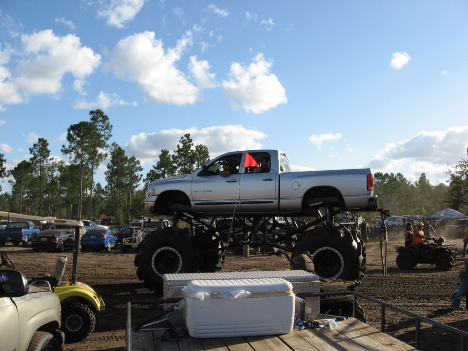 Silver pickup truck body on monster truck chassis with large tires, parked outdoors with other vehicles and trees in background