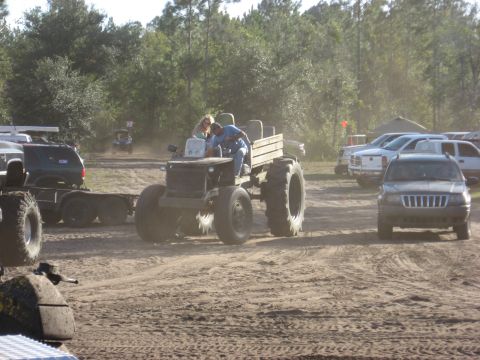 1994 Ford Bronco - A custom-built tractor with large tires and a wooden bed is seen with two people on board, surrounded by other vehicles in a dusty outdoor…