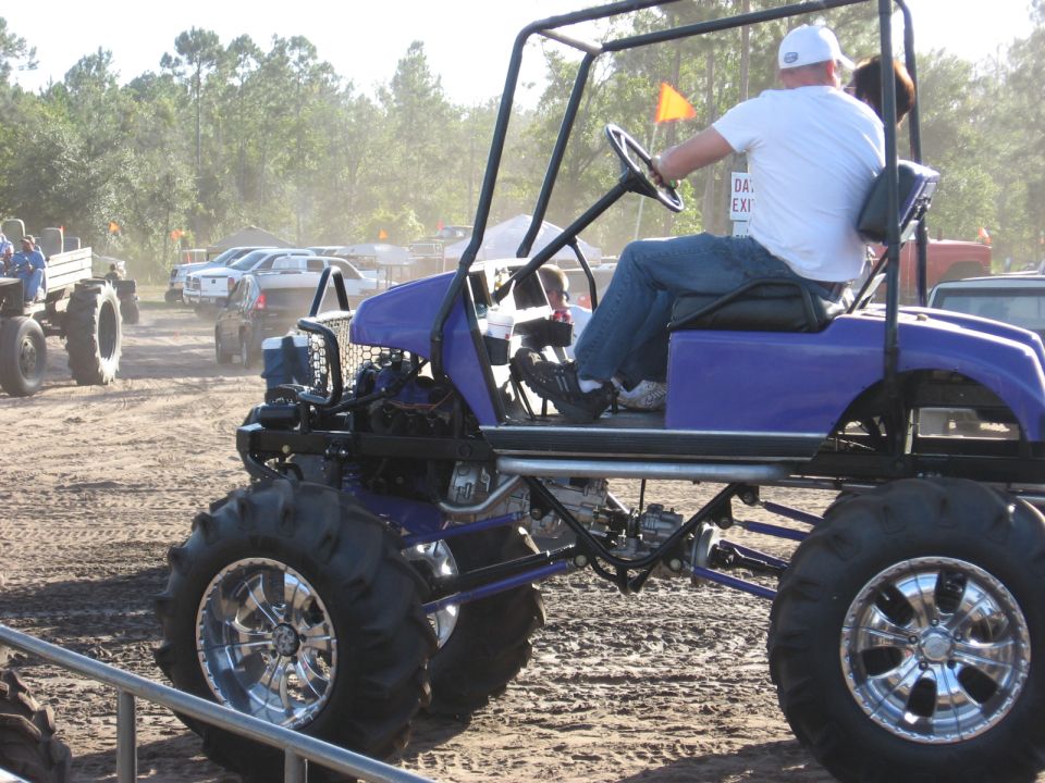 Purple custom golf cart with oversized tires and chrome rims, driven by a man in a white shirt and cap