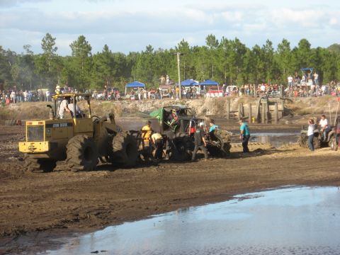 1994 Ford Bronco - A muddy scene unfolds at a mud bogging event, a John Deere tractor and several modified off-road vehicles being worked on by people.
