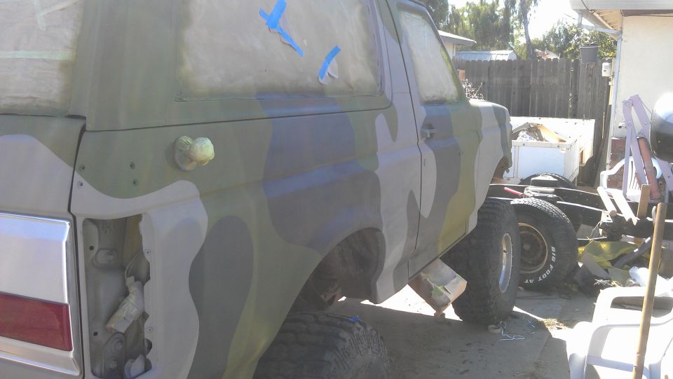 Ford Bronco with camouflage paint job, rear quarter panel removed, sitting on blocks