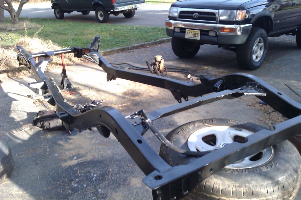 Black vehicle frame on the ground with a spare tire underneath. Two trucks are visible in the background