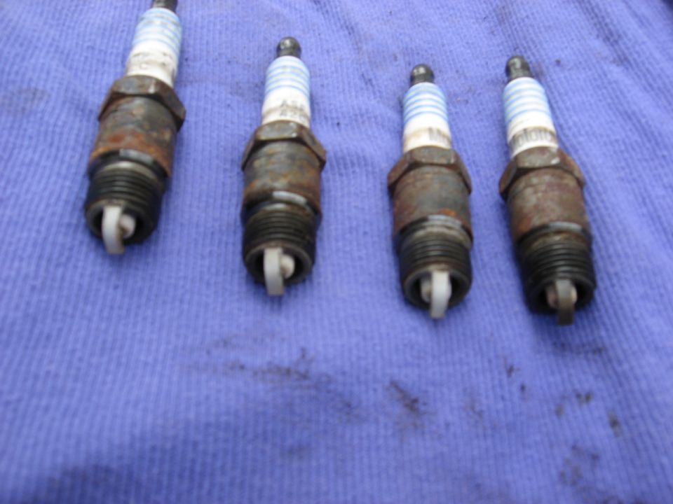 Four used spark plugs laid out on a blue cloth. Some have rust on their metal casings
