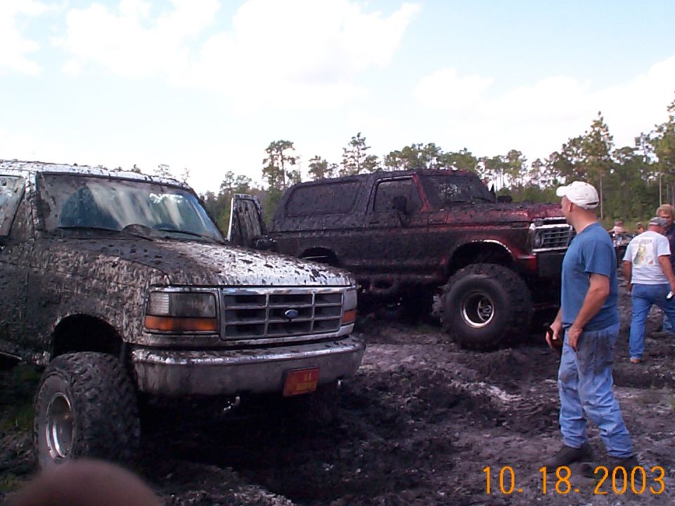 Two mud-covered lifted Ford trucks at a mud bog, man watching at right