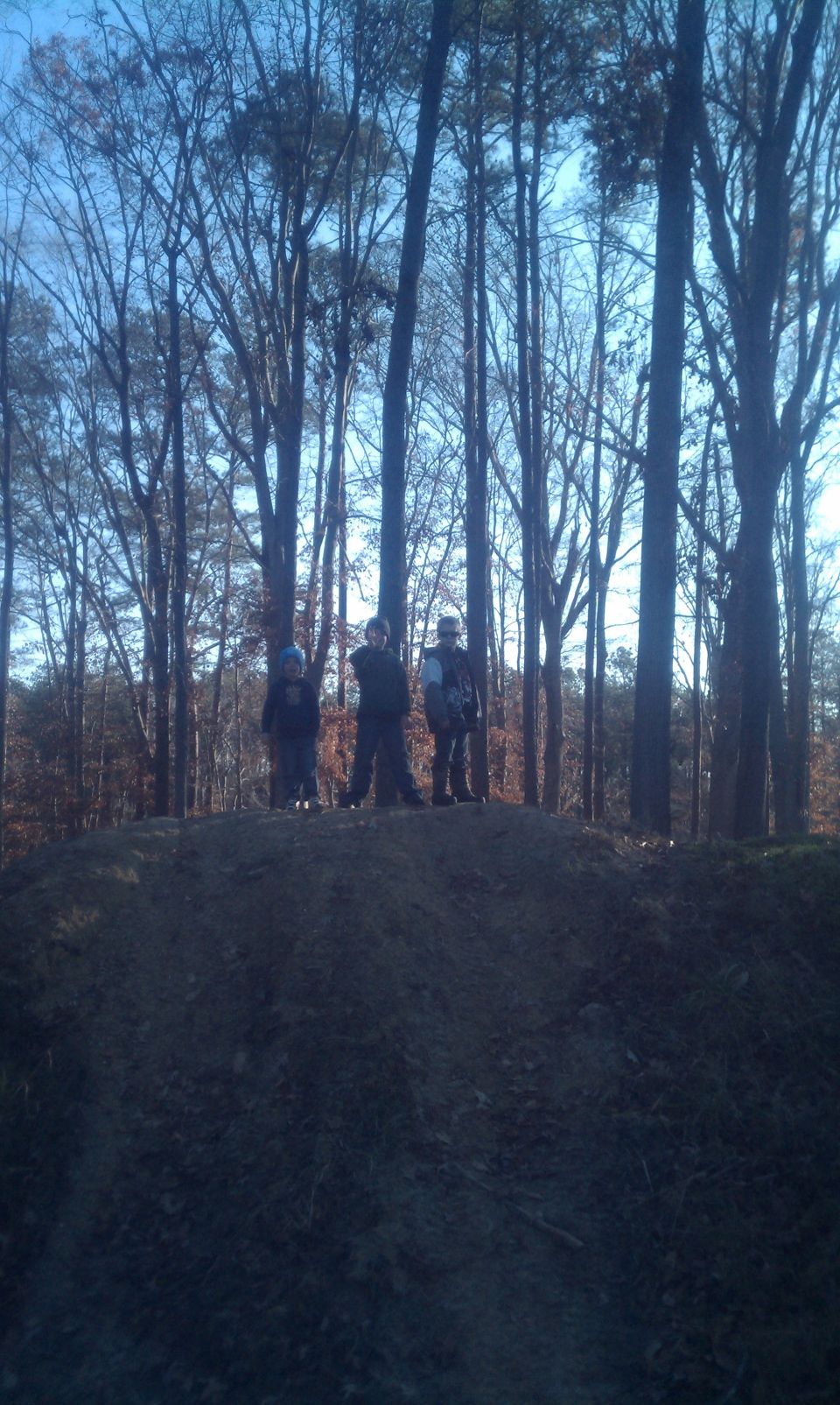 Three people stand on a dirt mound in a wooded area. A 'SUPERMOTORS' watermark is visible at the bottom